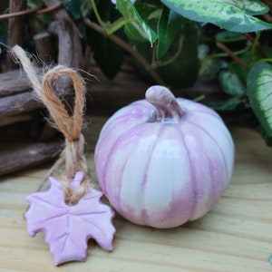 May include: A small, white ceramic pumpkin with a light purple glaze. The pumpkin has a stem and is sitting on a wooden surface. There is a small, purple ceramic leaf next to the pumpkin.
