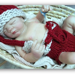 May include: A sleeping baby doll wearing a red and white knitted hat and matching red knitted diaper. The doll is lying in a wicker basket.