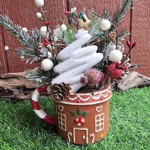 May include: A gingerbread house shaped mug with a white candy cane handle. The mug is filled with a white ceramic Christmas tree, red and white berries, and pine sprigs.