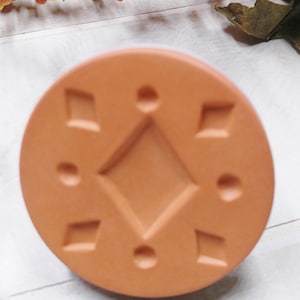May include: A terracotta-colored round coaster with a raised geometric design. The design features a large diamond shape in the center surrounded by smaller diamonds and circles.