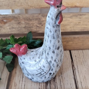 Puede incluir: Una figurita de pollo de cer&aacute;mica blanca con manchas negras y una cresta y barbillas rojas. El pollo est&aacute; de pie sobre una superficie de madera con una regla para la escala. El pollo mide aproximadamente 7,5 cm de alto.