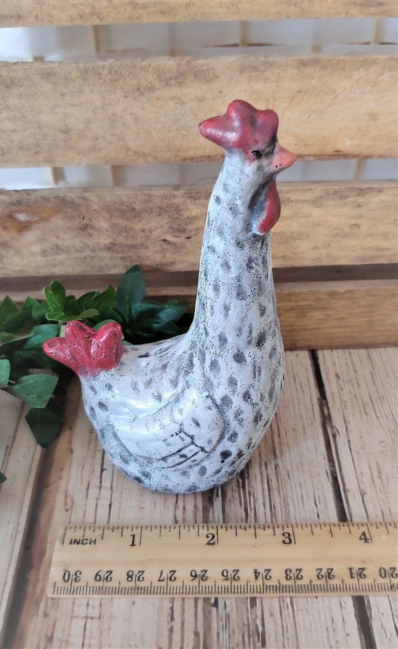 Puede incluir: Una figurita de pollo de cer&aacute;mica blanca con manchas negras y una cresta y barbillas rojas. El pollo est&aacute; de pie sobre una superficie de madera con una regla para la escala. El pollo mide aproximadamente 7,5 cm de alto.