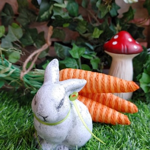 May include: A white ceramic bunny rabbit figurine with a green ribbon around its neck, sitting on green grass with three orange carrots behind it. A red and white mushroom is in the background.