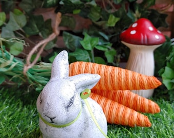 May include: A white ceramic bunny rabbit figurine with a green ribbon around its neck, sitting on green grass with three orange carrots behind it. A red and white mushroom is in the background.