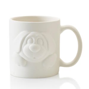 May include: White ceramic mug featuring a raised relief of a smiling dog's face. The mug has a rounded handle and a smooth, matte finish. The dog's features include large, floppy ears and a prominent nose.