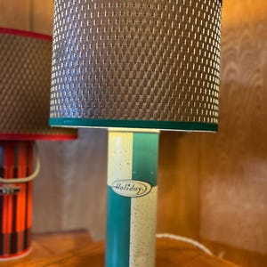 Könnte beinhalten: Eine Vintage-Lampe in Grün und Weiß mit einem braunen Korbgeflecht-Schirm. Der Lampenfuß trägt den Schriftzug "Holiday".