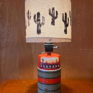Könnte beinhalten: Eine einzigartige Tischlampe mit einem zylindrischen Lampenschirm mit schwarzen Kaktussilhouetten. Der Sockel ist ein wiederverwendeter orangefarbener Behälter mit dem Aufkleber "Saguaro National Park". Die Lampe hat einen roten Streifen und einen Metallsockel.