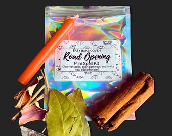 May include: A holographic pouch labeled Easy Bake Coven Road Opening Mini Spell Kit with text about clearing obstacles. It includes an orange taper candle, dried bay leaves, a small clear crystal, and two cinnamon sticks arranged in front.