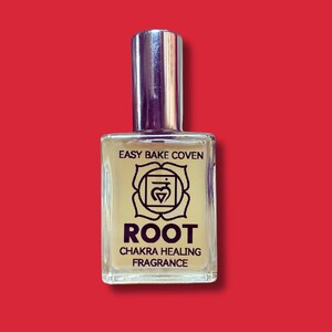 Op de afbeelding: Een heldere glazen fles van "Root Chakra Healing Fragrance" met een zilveren dop. Het etiket bevat de tekst "Easy Bake Coven" boven een symbool en het woord "ROOT". De achtergrond is effen rood.
