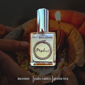 Puede incluir: Un frasco de perfume de vidrio transparente con tapa plateada, etiquetado como "Easy Bake Coven" y "Orphic Fragrance Potion". La etiqueta presenta un diseño de serpiente. El frasco se sostiene en manos ahuecadas, con las palabras "incienso", "palo santo" y "té verde" en la parte inferior.