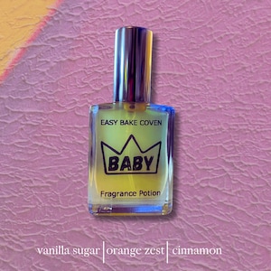 May include: A perfume bottle with a silver cap and a clear glass body. The label reads "EASY BAKE COVEN BABY Fragrance Potion." The bottle has a yellow and clear liquid inside. The bottom of the image lists "vanilla sugar | orange zest | cinnamon."