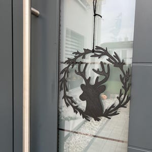 Könnte beinhalten: Ein schwarzer Metall-Rentierkopf-Kranz mit einer schwarzen Schnur, die an einem Haken an einer grauen Tür hängt.