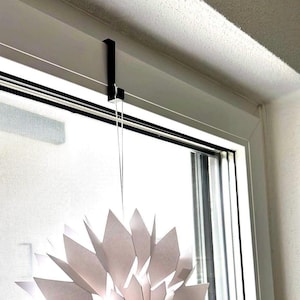 Peut inclure: Une décoration en forme de flocon de neige en papier blanc suspendue à un crochet en métal noir fixé à un cadre de fenêtre blanc.