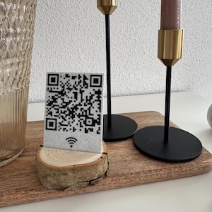 Könnte beinhalten: Eine QR-Code-Karte mit einem Wi-Fi-Symbol, die auf einem Holzsockel präsentiert wird, liegt auf einem Holztablett. Zwei schwarze Kerzenhalter mit goldenen Akzenten und rosa Kerzen befinden sich im Hintergrund.