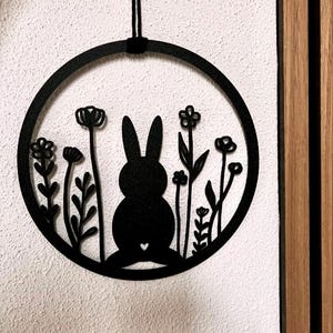 Könnte beinhalten: Schwarzer Metallring mit einer Silhouette eines Kaninchens und Blumen darin. Der Ring hängt an einer schwarzen Schnur.