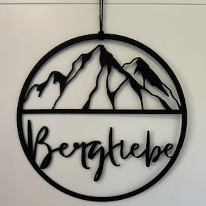 Könnte beinhalten: Schwarzer Holz-Kreis mit einer Bergsilhouette und dem Wort "Bergliebe" in einer kursivschrift.