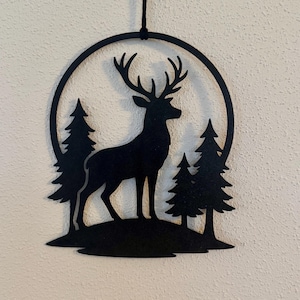 Könnte beinhalten: Schwarze Metall-Wandkunst mit einer Hirschsilhouette in einem kreisförmigen Rahmen. Der Hirsch steht inmitten stilisierter Kiefern und schafft eine Waldszene. Das Kunstwerk wird von einer schwarzen Schnur aufgehängt, geeignet für die Inneneinrichtung.