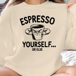 May include: A cream-coloured sweatshirt with the text "ESPRESSO YOURSELF... OR ELSE" in black. The graphic features an angry coffee cup with arms. The sweatshirt is a casual, comfortable style.