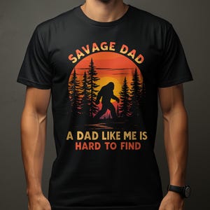 May include: Black t-shirt with a Bigfoot graphic in front of a sunset scene. Text reads "SAVAGE DAD" above and "A DAD LIKE ME IS HARD TO FIND" below the image. The shirt is worn by a person.