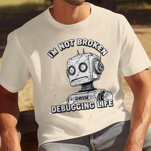 Könnte beinhalten: Cremefarbenes T-Shirt mit einem Roboter-Grafik und dem Text "I'M NOT BROKEN" darüber und "JUST DEBUGGING LIFE" darunter. Der Roboter hat einen quadratischen Kopf und kreisförmige Augen. Das T-Shirt wird von einer Person getragen.