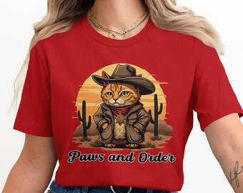 May include: Red t-shirt with a cartoon illustration of a cat wearing a cowboy hat and holding a badge. The text "Paws and Order" is printed below the image.
