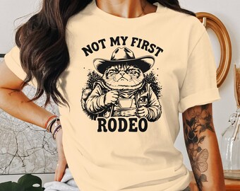 Pode incluir: Uma camiseta amarela clara com uma ilustração em preto e branco de um gato usando um chapéu de cowboy e segurando uma xícara de café. O texto "NOT MY FIRST RODEO" está impresso na camiseta.