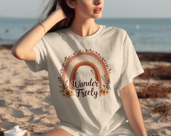 Puede incluir: Camiseta color crema con un diseño de arcoíris en acuarela en tonos marrón, naranja y amarillo, con la frase "Wander Freely". El arcoíris está rodeado de elementos florales. Prenda de vestir informal.