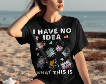 以下が含まれることがあります： 「I HAVE NO IDEA WHAT THIS IS」の文字と、ルービックキューブ、フロッピーディスク、カセットテープ、携帯ゲーム機などのレトロなグラフィックがプリントされた黒いTシャツ。