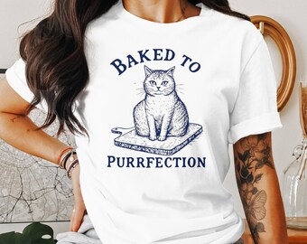 May include: White t-shirt with a blue graphic of a cat sitting on a cutting board. The text "Baked to Purrfection" is printed below the cat.