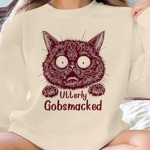 May include: Cream-colored sweatshirt featuring a maroon illustration of a surprised cat face with large eyes and the text "Utterly Gobsmacked" below. The cat's expression is one of shock. The sweatshirt is a casual, long-sleeved garment.