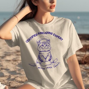 May include: White t-shirt with a blue graphic of a cat wearing a chef's hat, kneading dough. Text above the cat reads "CERTIFIED KNEADING EXPERT", and below it, "MEOW MADE". The shirt is a casual, short-sleeved design.