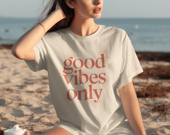 May include: A white t-shirt with the words "good vibes only" in a coral-colored, stylized font. The design includes sunburst graphics. The shirt is worn by a person sitting on a sandy beach.