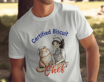Puede incluir: Camiseta blanca con un gráfico azul de una mujer con un gato horneando galletas. El texto "Certified Biscuit Chef" está impreso en azul en la camiseta.