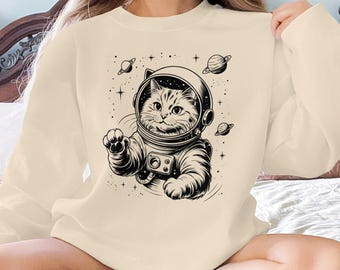 May include: Cream-colored sweatshirt featuring a black and white illustration of a cat in an astronaut helmet and spacesuit. The design includes planets and stars, creating a space-themed graphic. The sweatshirt is a casual, long-sleeved garment.