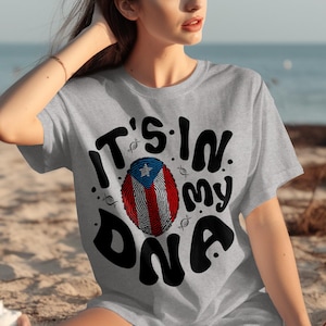 Puede incluir: Camiseta gris claro con el texto "IT'S IN MY DNA" en negro, con un diseño de huella dactilar con los colores de la bandera puertorriqueña. La camiseta es de manga corta.