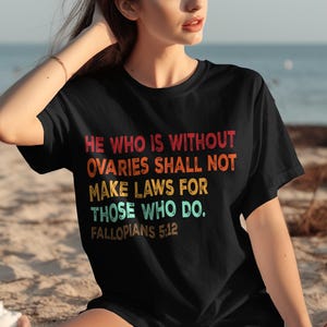 May include: Black t-shirt with the text "HE WHO IS WITHOUT OVARIES SHALL NOT MAKE LAWS FOR THOSE WHO DO. FALLOPIANS 5:12" in red, orange, yellow, and blue lettering. The shirt is worn by a person on a beach.