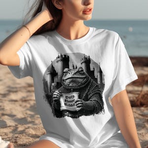 May include: White t-shirt featuring a black and white illustration of a frog in armor holding a scroll that reads "Duty, Honor, Ribbit" with a castle backdrop. The shirt is a casual, short-sleeved style.