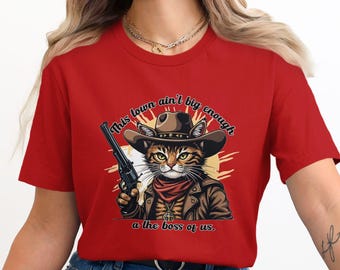 Peut inclure: T-shirt rouge avec un motif représentant un chat portant un chapeau de cowboy et tenant un pistolet. Le texte sur le t-shirt est "This town ain't big enough for the boss of us."