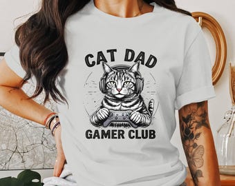 Puede incluir: Una camiseta blanca con una ilustración en blanco y negro de un gato con auriculares y sosteniendo un mando de videojuego. El texto "Cat Dad Gamer Club" está impreso en la camiseta.