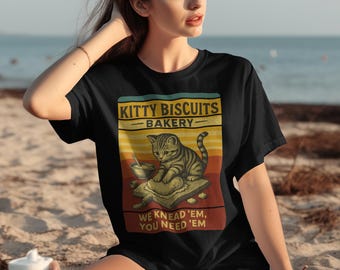 Może przedstawiać: Czarny t-shirt z grafiką w stylu retro przedstawiającą kota pręgowanego ugniatającego ciasto, z napisem "Kitty Biscuits Bakery" i "We Knead 'em, You Need 'em". Projekt ma schemat kolorów w stylu vintage.
