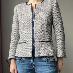 Puede incluir: Una chaqueta de tweed blanca y negra con ribete negro y cuello en V. La chaqueta se lleva sobre unos vaqueros azules.