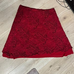 May include: A red skirt with a floral pattern. The skirt has a layered design, with a darker floral overlay over a solid red underlayer. The skirt is made of a lightweight material and has a flared shape.