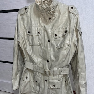 Puede incluir: Chaqueta beige claro con un patrón texturizado, cintura con cinturón, múltiples bolsillos y cierres de botones. La chaqueta tiene un cuello alto y charreteras, con la marca "Barbour" visible en la parte delantera.