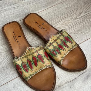 Könnte beinhalten: Ein Paar braune Ledersandalen mit einem roten und grünen Chili-Pfeffer-Druck. Die Sandalen haben ein schlichtes Design und werden von der Marke "Muxart Barcelona" hergestellt.