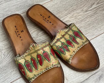 May include: A pair of brown leather sandals with a red and green chili pepper print. The sandals have a simple design and are made by the brand "Muxart Barcelona".