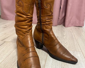 Può includere: Un paio di stivali da cowboy in pelle marrone con punta a punta e una cerniera laterale. Gli stivali hanno un design decorativo sulla parte superiore del gambale.