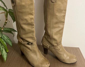 May include: A pair of tan leather knee-high boots with decorative stitching and a buckle detail. The boots have a block heel and a pointed toe.