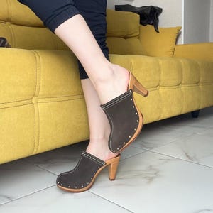 Può includere: Zoccoli in pelle scamosciata marrone con tacchi e suole in legno. Gli zoccoli hanno la punta arrotondata e sono impreziositi da piccole borchie argentate e cuciture bianche. I tacchi sono alti circa 7,6 cm.