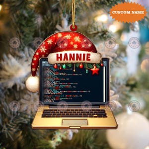 May include: A Christmas ornament shaped like a laptop, with a Santa hat design. The screen displays code, and the name "HANNIE" is printed on the hat. The ornament is gold and red, with a gold string for hanging.