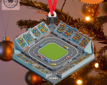 May include: A 2D flat Christmas ornament depicting a race track with cars, spectator stands, and a green infield. The ornament has a red ribbon for hanging and the text "CUSTOM NAME" at the top. The name "HENRY" is printed at the bottom.